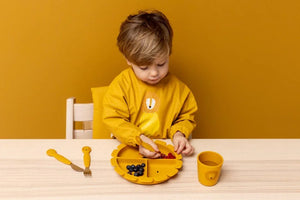 Tout-petit utilisant un set de repas en silicone jaune moutarde motif lion, incluant gobelet, assiette et couverts