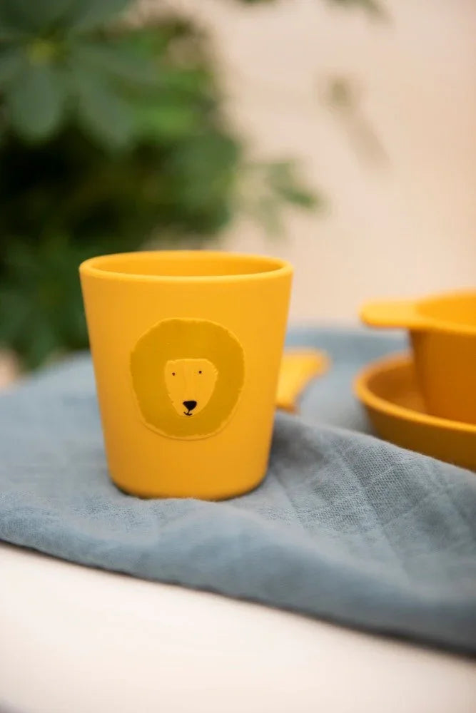 Gobelet enfant en silicone jaune avec motif tête de lion, posé sur un tissu bleu