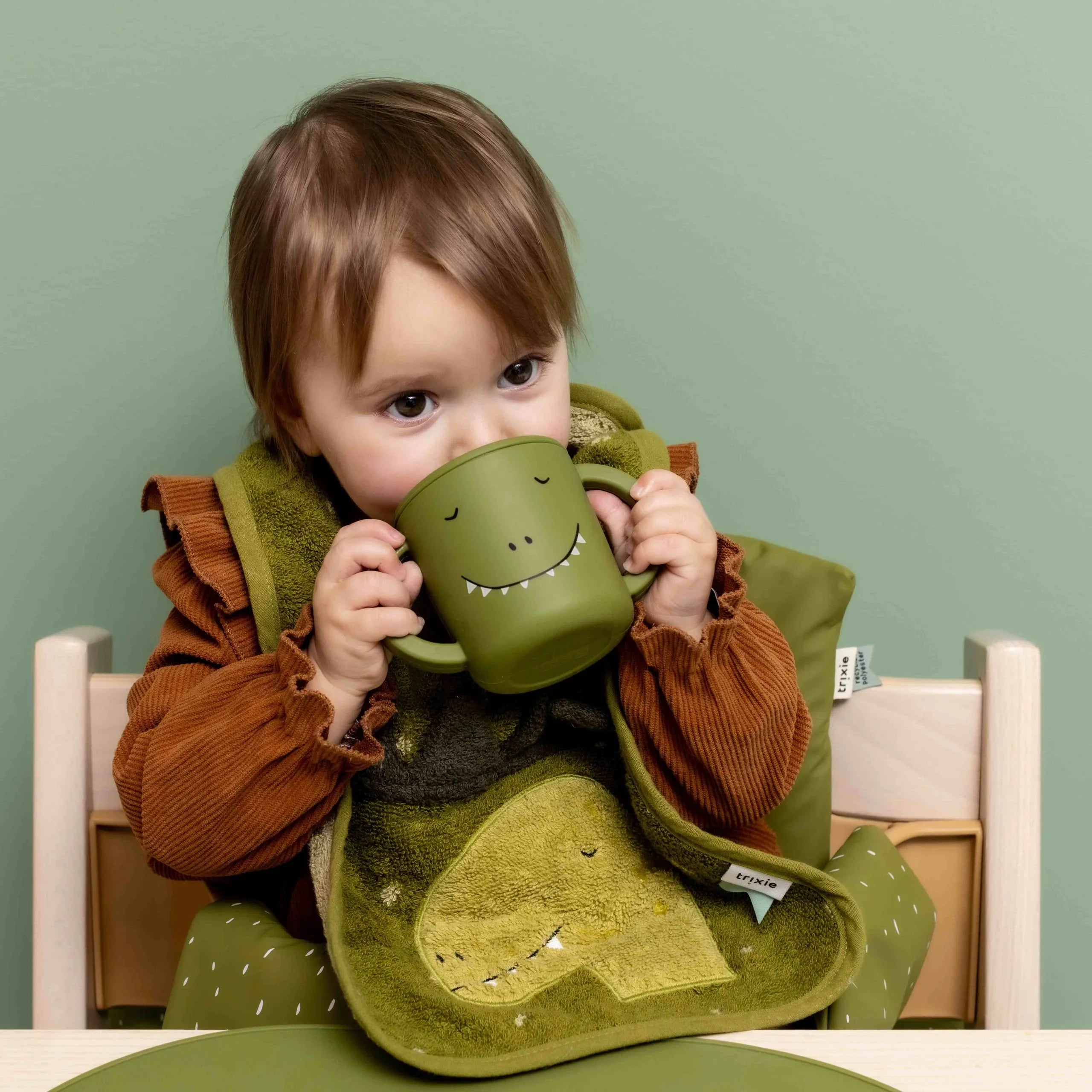 Niño pequeño usando un vaso de aprendizaje de silicona verde con diseño de dinosaurio, con babero y mantel individual a juego