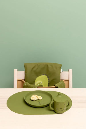 green silicone sippy cup with dinosaur face, matching plate, and placemat on light wood table