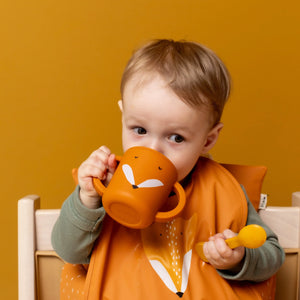 Tout-petit utilisant un gobelet à bec en silicone orange motif renard, avec bavoir renard et cuillère jaune assortis