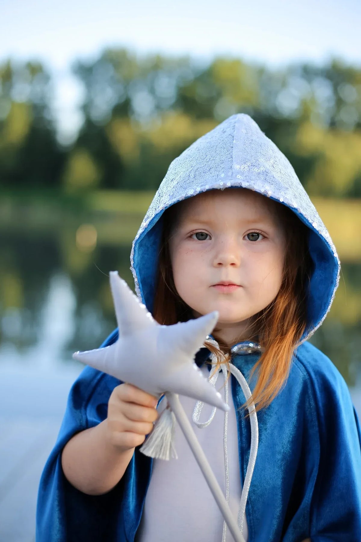 Child wearing Snow Queen cape with sparkling silver sequin hood and holding a silver star wand beside a lake