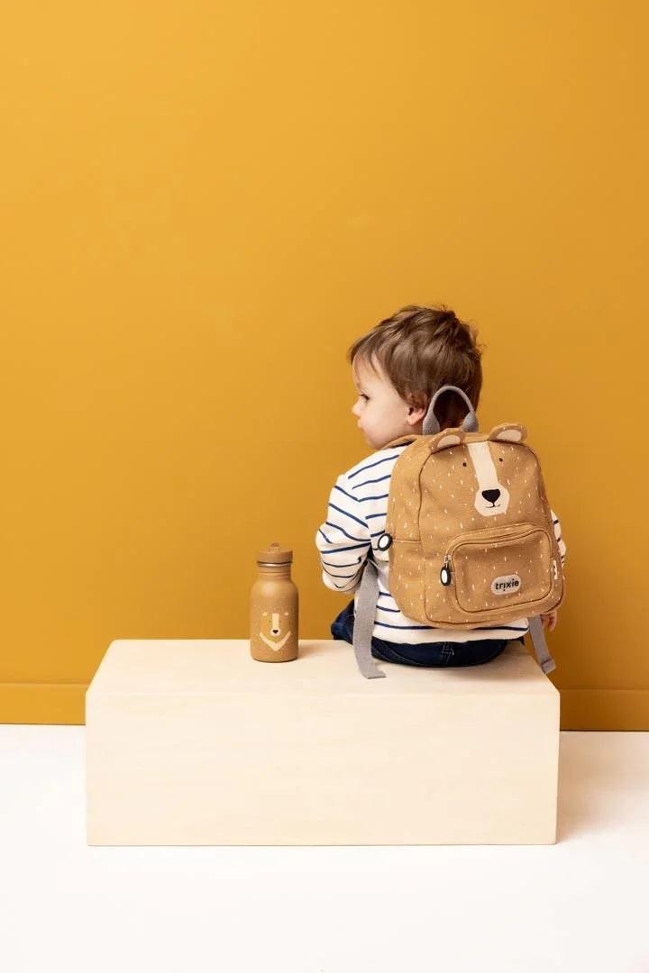 Kind mit braunem Bären-Rucksack und passender Edelstahl-Wasserflasche auf einer Bank vor senfgelbem Hintergrund
