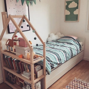 Natural house-shaped shelf used beside a child’s bed with books and a wooden dollhouse
