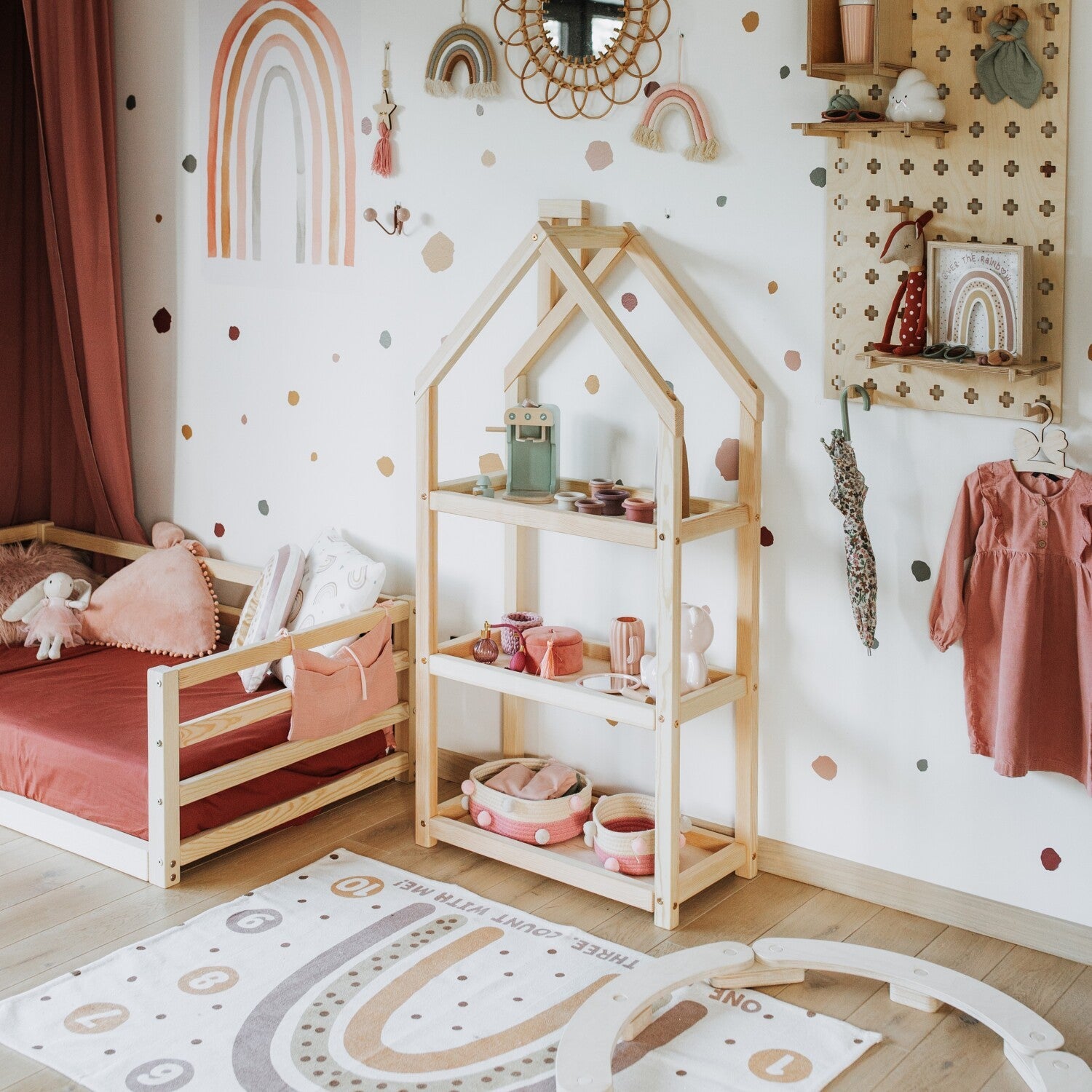 Natural pine house-shaped kids shelf styled beside a low wooden bed in a pastel rainbow children’s room