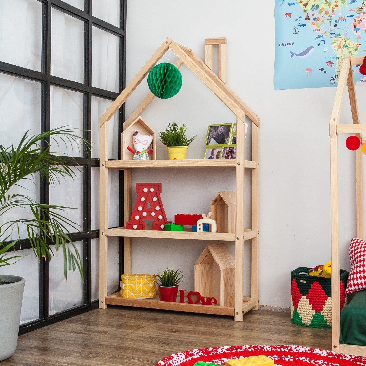 Natural wooden house-shaped shelf styled with toys, plants and family photographs beside a children’s bed