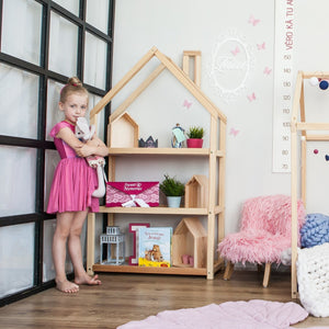 Natural house-shaped shelf beside a child in a pink dress, filled with books and decorative storage