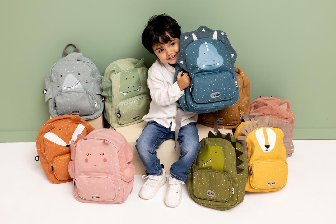 Child sitting among colourful Trixie animal backpacks while holding a blue dinosaur backpack