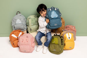 Child sitting among colourful Trixie animal backpacks while holding a blue dinosaur backpack