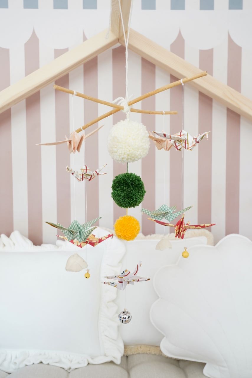 Moi Mili Vintage Vibe origami mobile with patterned paper cranes and colourful pompoms above white nursery cushions