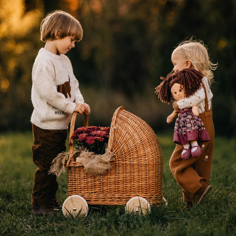 Bambini che giocano all'aperto con una carrozzina in vimini naturale riempita di fiori e una bambola di pezza