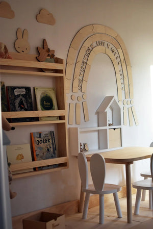 Decorazione da parete arcobaleno in legno con scritta incisa, libreria in legno chiaro per cameretta e mobili bianchi per bambini in una stanza giochi accogliente